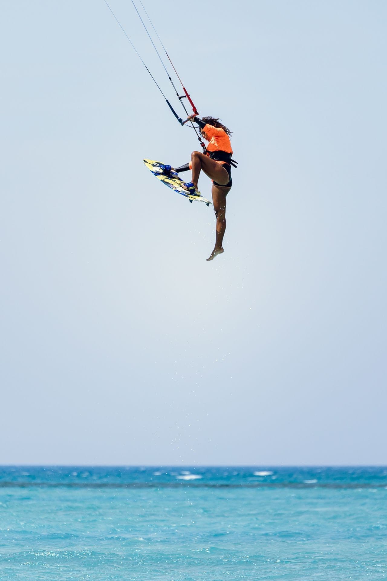 A kitesurfer in an orange top inverted mid-jump, holding the board, high above open water