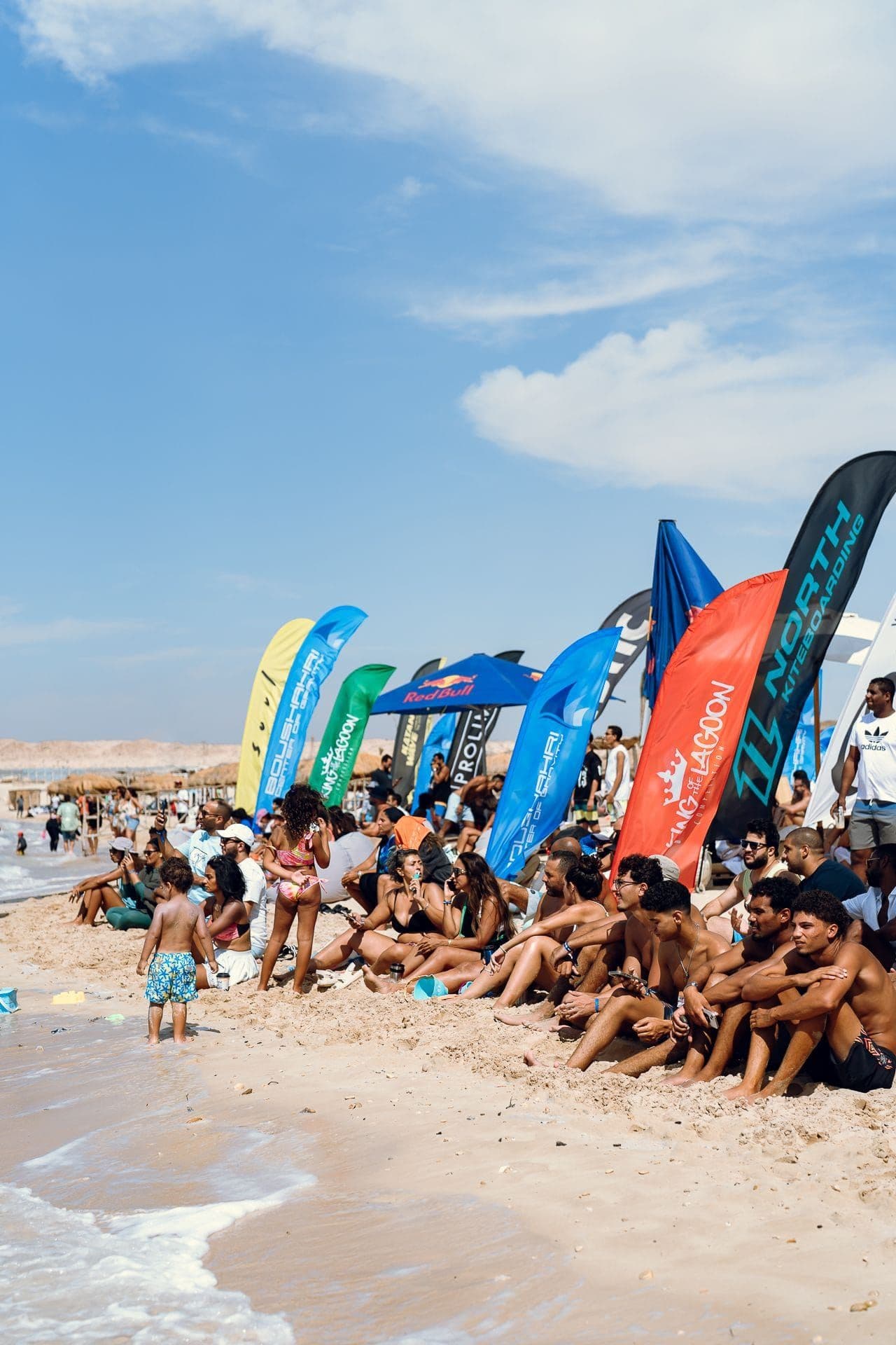 Spectators sitting along the waterline beneath a row of event banners, a small child standing at the edge of the shallows