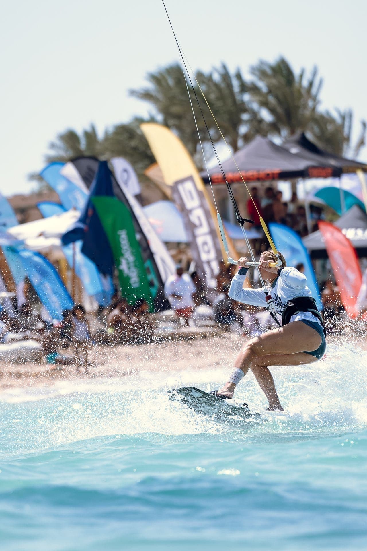 A kitesurfer carving through spray close to shore, with event flags and spectators blurred behind