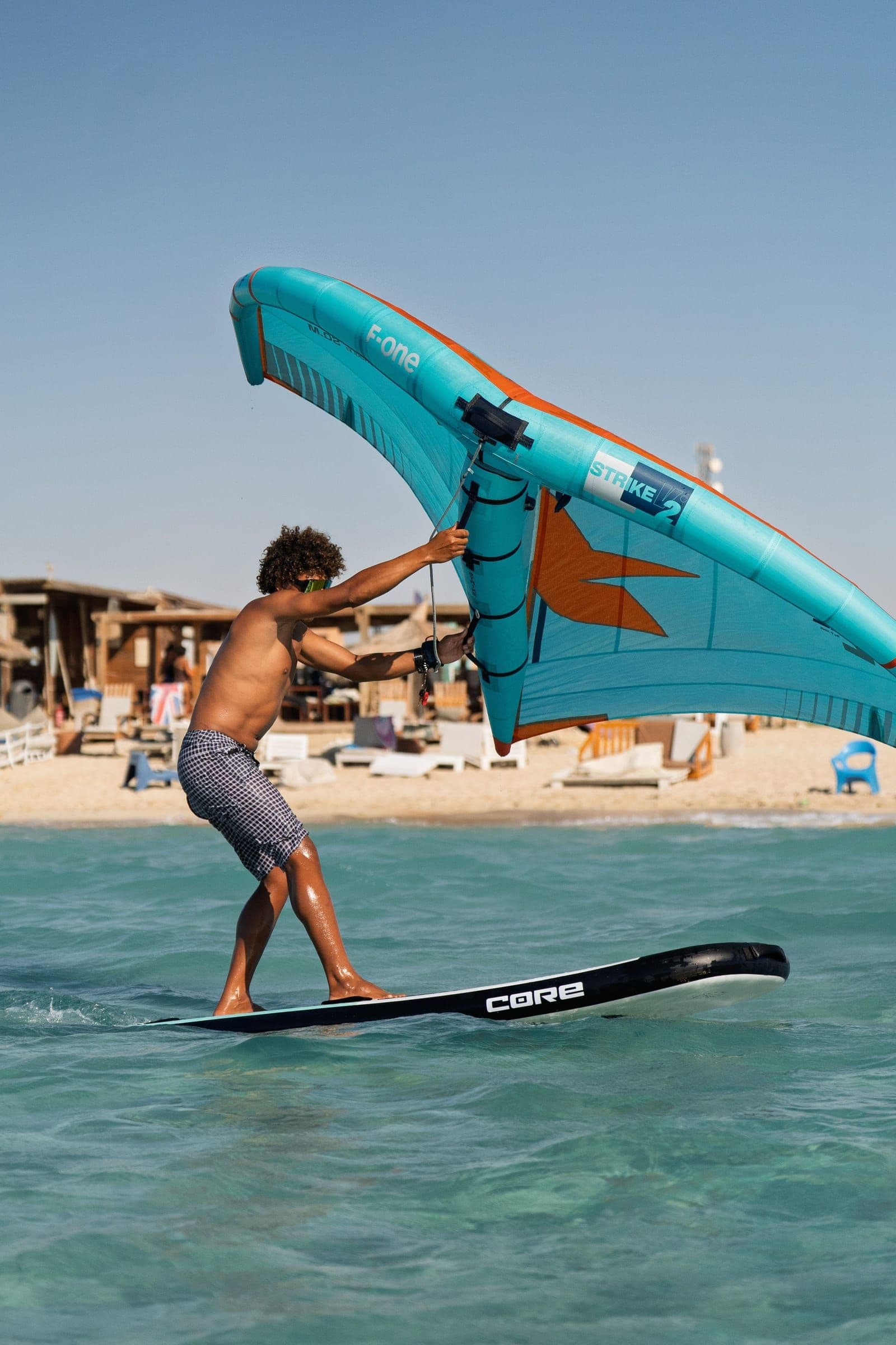 A rider crosses flat water on a wing board, with the beach club's loungers and pergola behind them.
