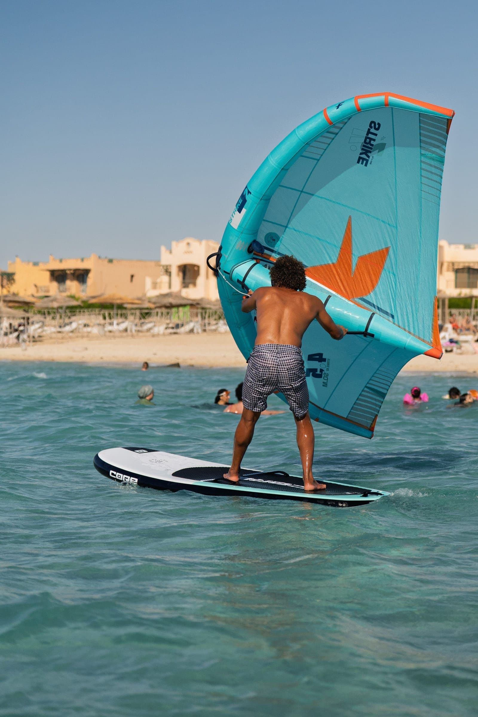A rider heads out across the lagoon under a turquoise wing, with people standing in the shallows behind.