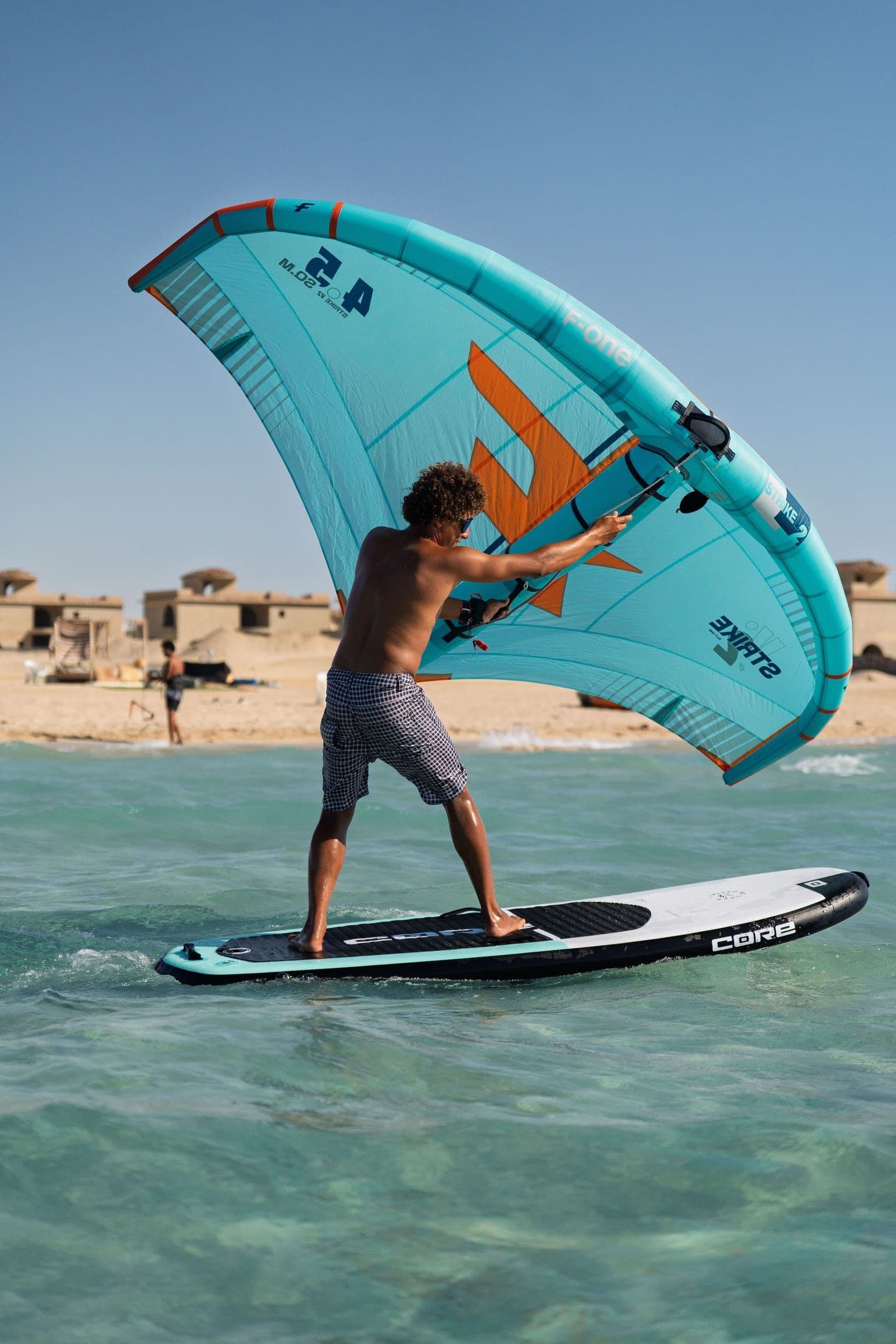 A rider stands on a wing board holding a turquoise wing, crossing clear shallow water in front of the beach.