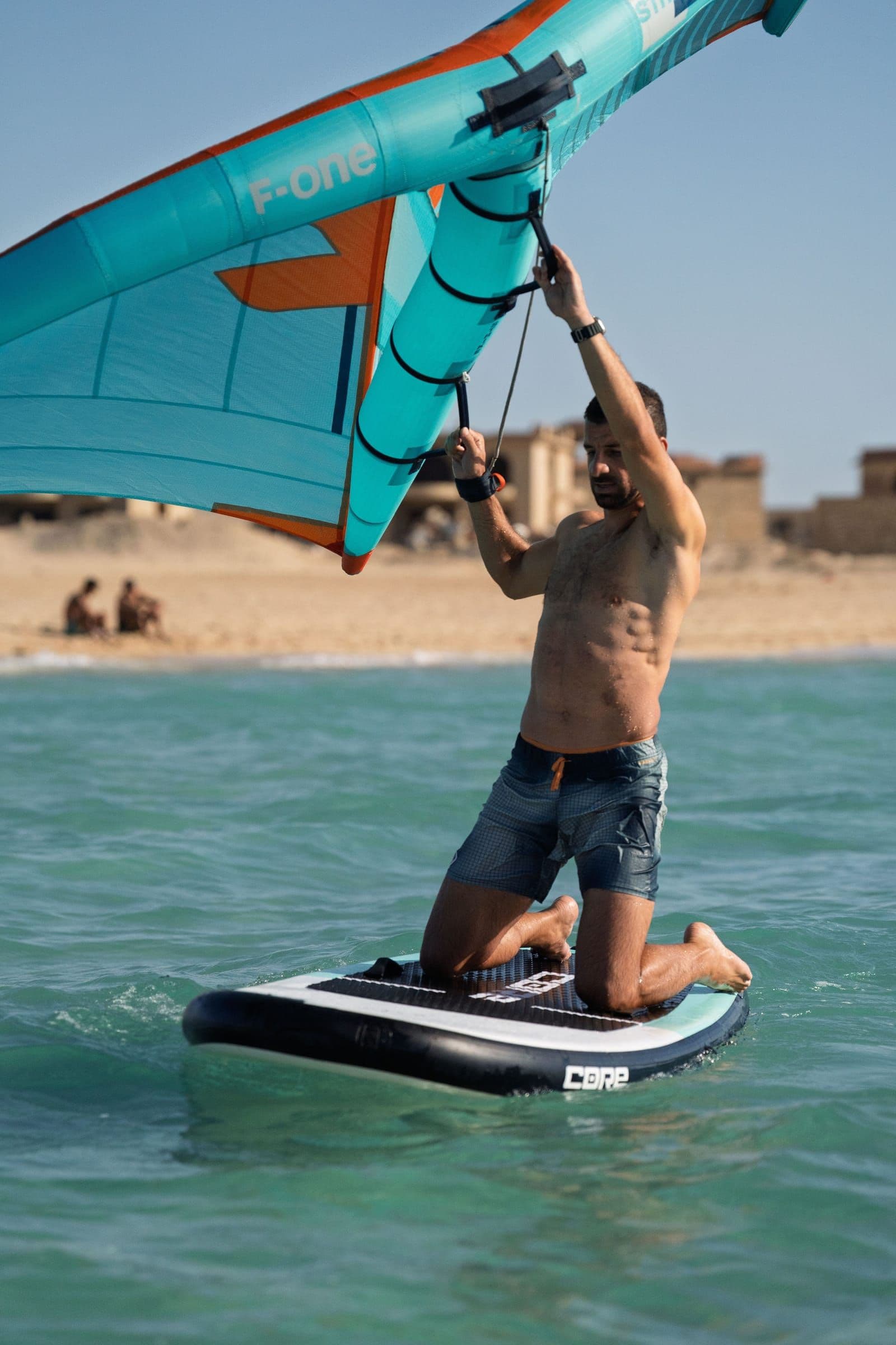 A rider kneels on a wing board with both hands on the wing's handles, holding it above their head.